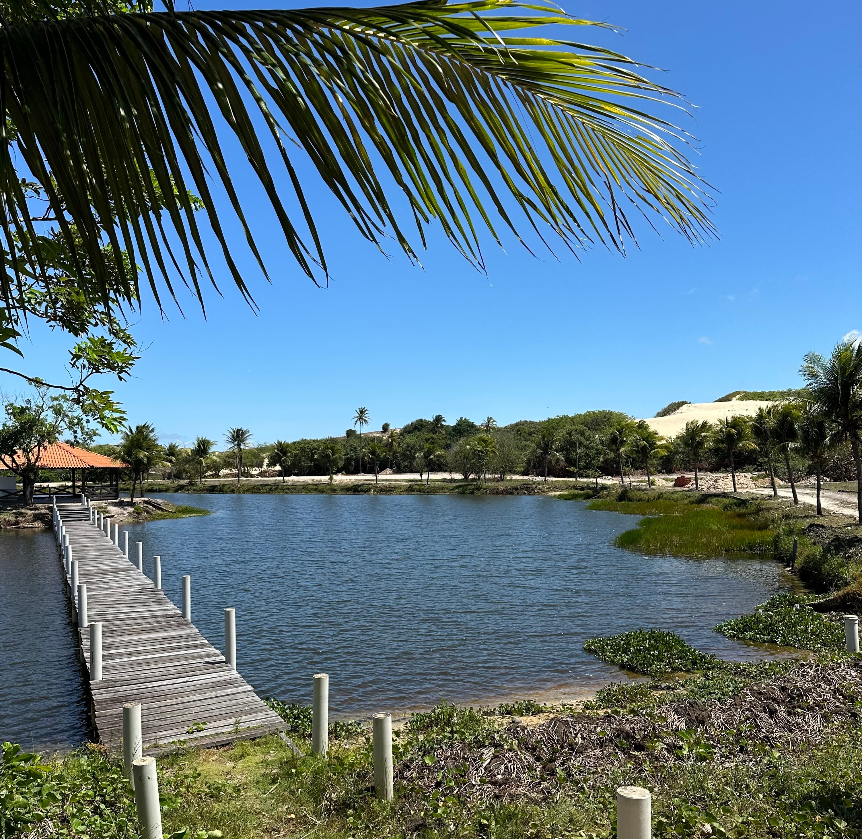 Lagoa natural do condomínio com passarela de madeira e dunas ao fundo.